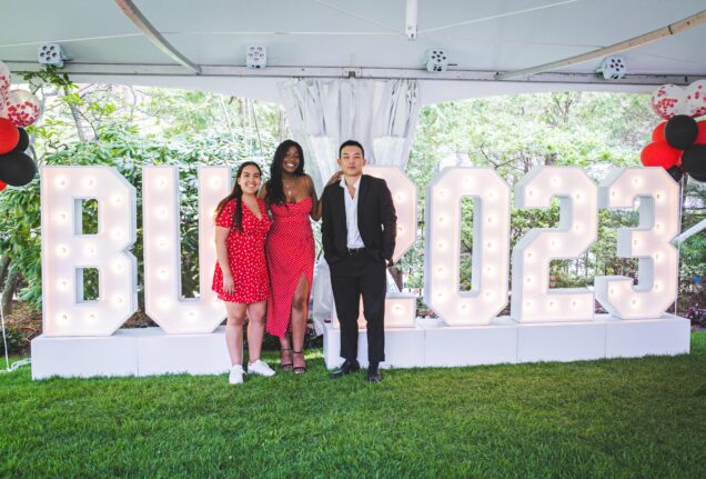 Jasmine Mijares (Questrom’23), Dumebi Onogwu (CAS’23), and Christian Hahm (Sargent’23) stand in front of a light bulb sign reading "BU 2023". The weather appears to be a bit blue and likely near the evening as it's very blue outside in the background beyond the tent limits. Photo by BU Photo.