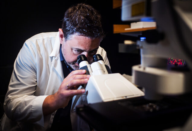 Dr. Basilis (Vasileios) Zikopoulos examines a sample by looking into a microscope. The image itself is tightly cropped but within a very dimly lit lab space. Photo by Jackie Ricciardi for Boston University Photography.
