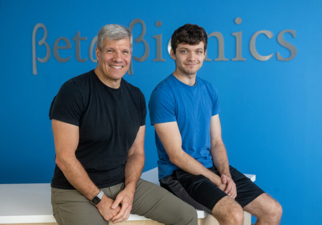 Ed Damiano with his son, David Damiano (CAS’21) who is Type One Diabetic and the inspiration behind Ed's work. They are sitting on a white reception desk situated in front of a sky blue colored wall adorning Beta Bionic's serif logotype.
