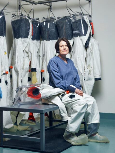 Elke Muhlberger, a professor in Microbiology & Director in Integrated Science Core poses sitting down in a room where lab techs switch out protective full-body suits. She looks exasperated, but hopeful. Photo by Doug Levy for Boston University Photography.