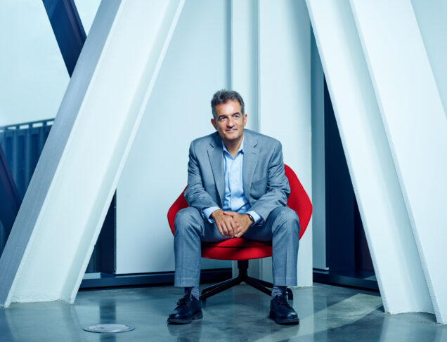 Yannis Paschalidis, Distinguished Professor of Engineering sits in a cool lighting temperature room on a red mid-century modern chair. His hair is a little dark with some grays of wisdom. He looks out towards the left of the camera. Photo by Doug Levy for Boston University.