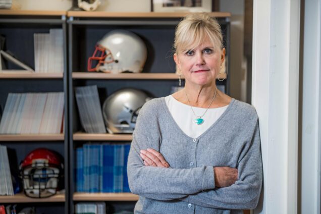Ann McKee, MD, Professor of Neurology & Pathology, Director, Neuropathology Core stands in an office which appears to have football helmets alongside scientific textbooks. She wears a powder gray cardigan with a turquoise necklace piece. She appears to be confidently pensive. Photo by Cydney Scott for Boston University Photography.