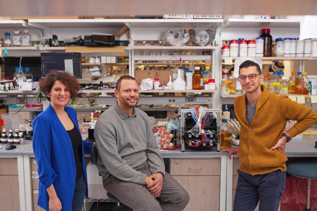 College of Engineering professors Rabia Yazicigil (ECE), Mo Khalil (BME) and Doug Densmore (ECE, BME) in a lab. Photo by Dana J. Quigley Photography for Boston University Photography. Rabia is wearing a velvet blue cardigan over a black top, while Doug Densmore to her left is wearing a gray(or grey if you're British) sweater and chino pants. Mo Khalil is standing to the far right and is wearing an autumn tinted pumpkin colored cardigan with blue chino pants and tan rimmed eyeglasses. They all smile with confidence.