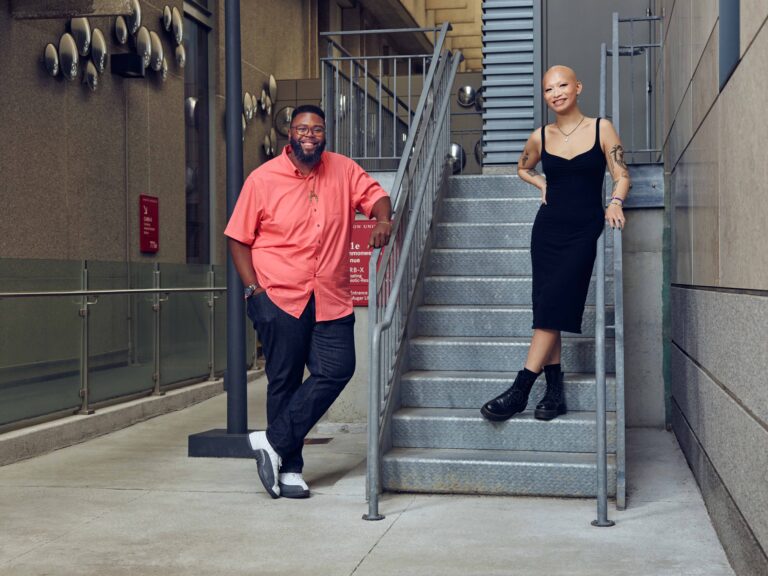 Dr. Anthony Jack is to the left while Katarina 'Kat' Quach(COM'24) stands to the right on an outdoor staircase posing along an industrial building on campus. They both have prideful smiles.