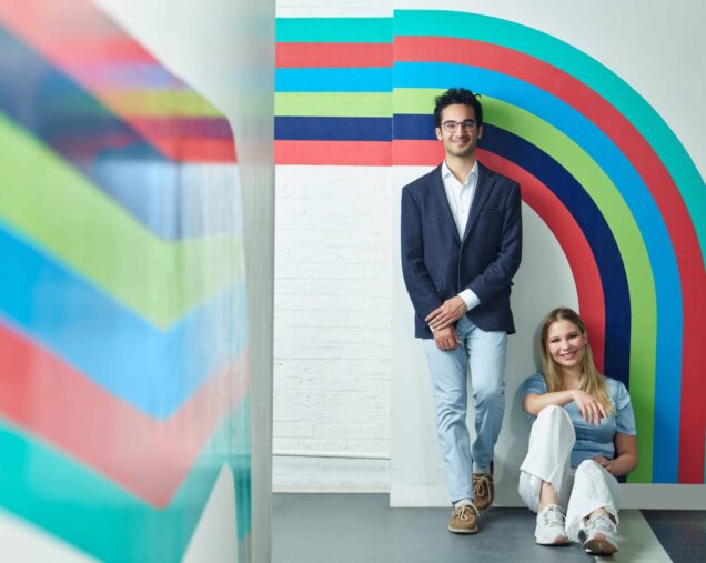 Caspian Chaharom(CAS'23) stands against a wall with Maria Gorsikikh(Questrom'23) sitting on the floor. They both share a confident smile, matched by a radiant pattern of wall art consisting of thick bold lines curving around the interior of the BUild Lab, with colors ranging from teals, blues, and lime to BU red.