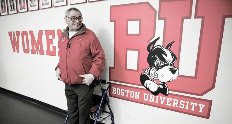 Older man, Elliot Driben, standing in front of a wall dedicated to women's athletics at a BU Women's Ice Hockey game