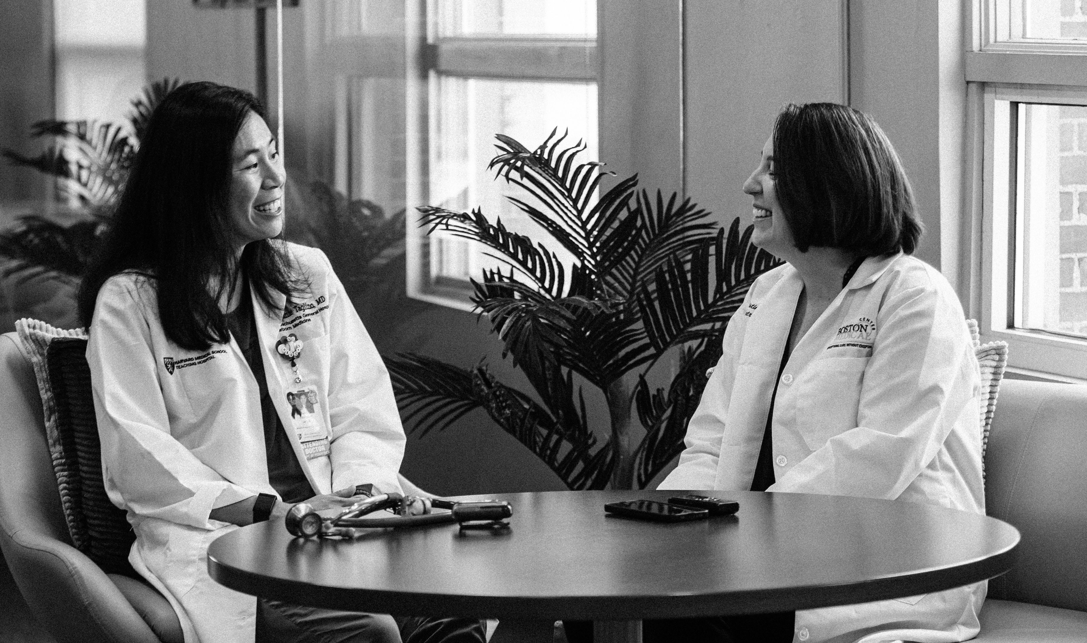 Two female doctors in lab coats sitting at a round table talking to one another