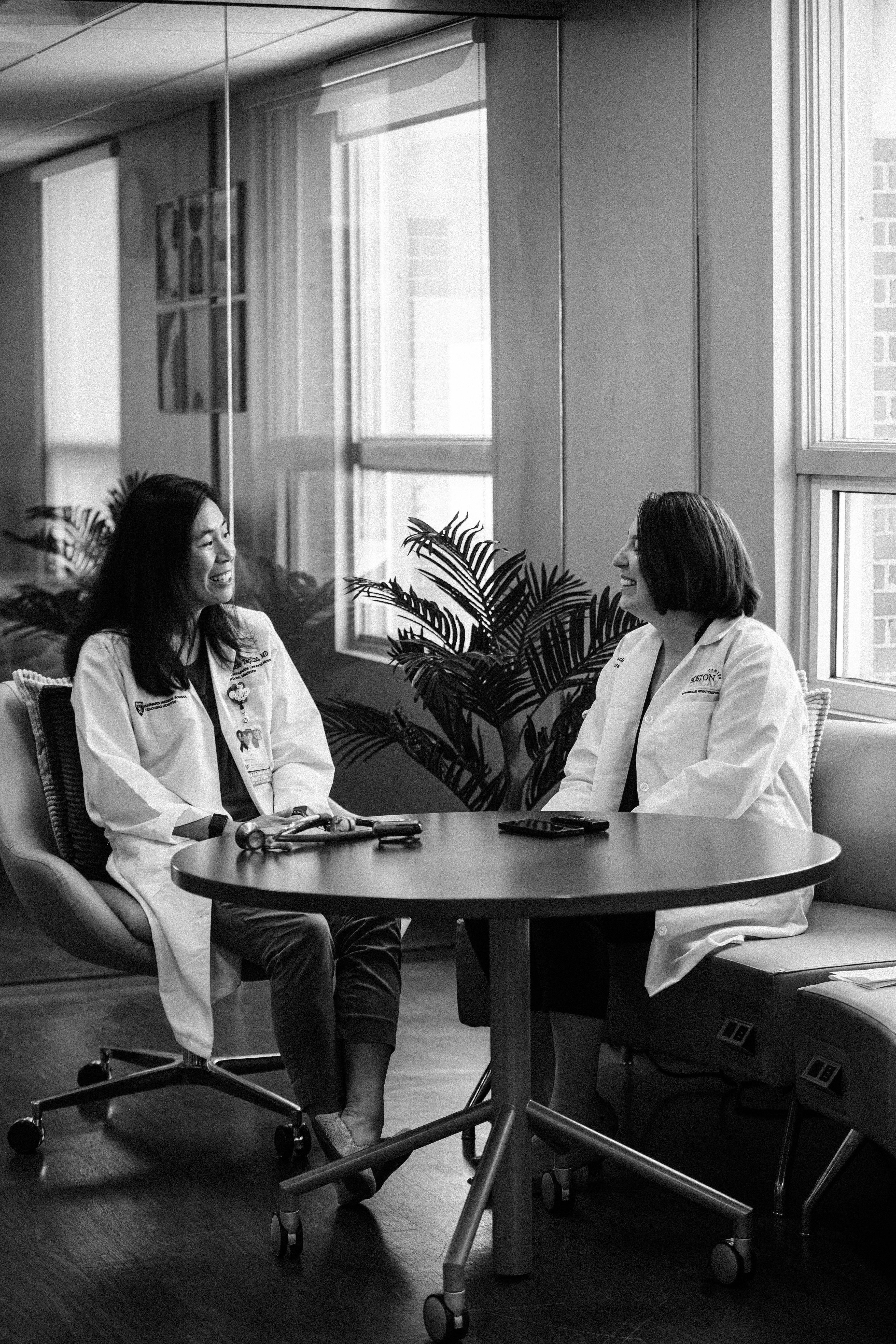 Two female doctors in lab coats sitting at a round table talking to one another