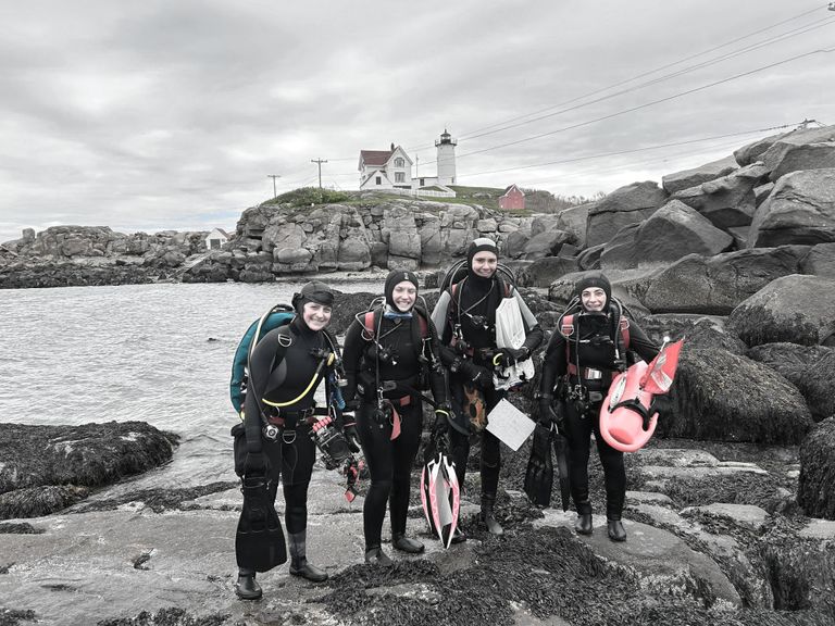 A group of four students wearing scuba gear in the shore of a body of water with a lighthouse behind them